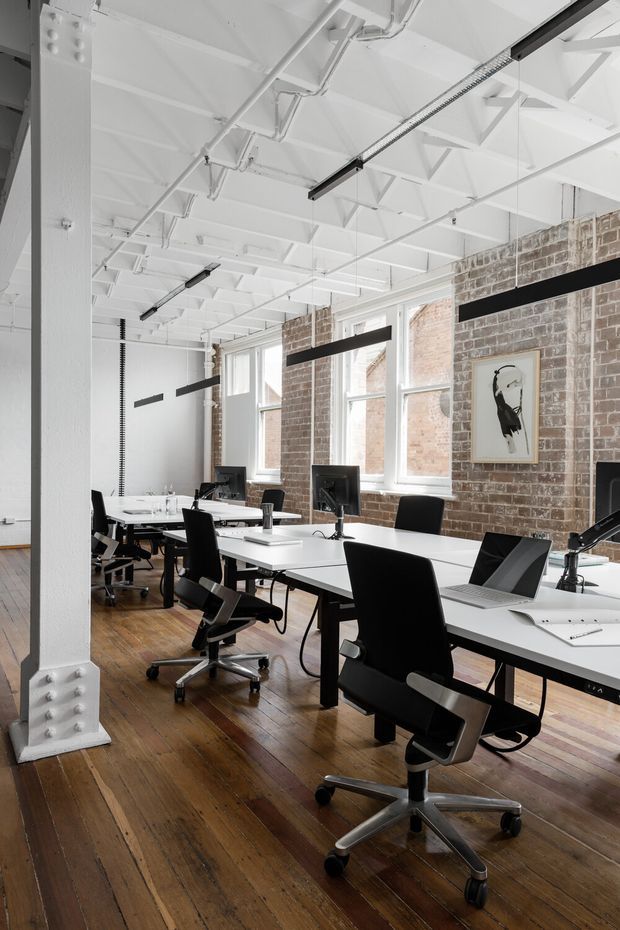 A bright open-plan commercial office with rows of white desks, black ergonomic task chairs, computer monitors, and laptops. The space features exposed white-painted structural beams and services, tall industrial-style windows, an exposed red-brick wall, suspended linear lighting, and aged timber floorboards.