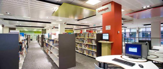 Bright modern library or information-services interior with rows of bookshelves, computer workstations, directional signage, colourful red and lime-green feature panels, a suspended acoustic grid ceiling, and grey carpet flooring.