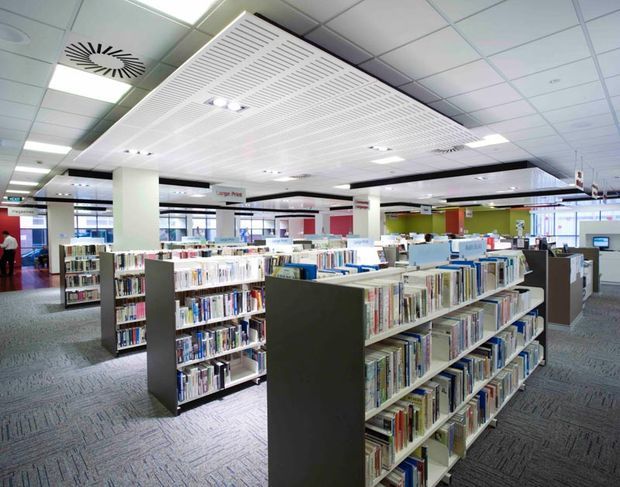 A bright, open-plan contemporary library interior with rows of low book shelving, white structural columns, carpeted flooring, extensive fluorescent lighting, and a suspended slatted ceiling feature. Reading and service areas are visible in the background.