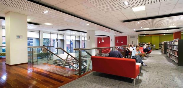 A bright contemporary public library interior viewed from an upper-level landing, with bookshelves, reading seating, a red upholstered bench, glass balustrades, a staircase, large windows, and a suspended grid ceiling. The space uses bold red and green feature walls alongside neutral carpet and polished timber flooring.