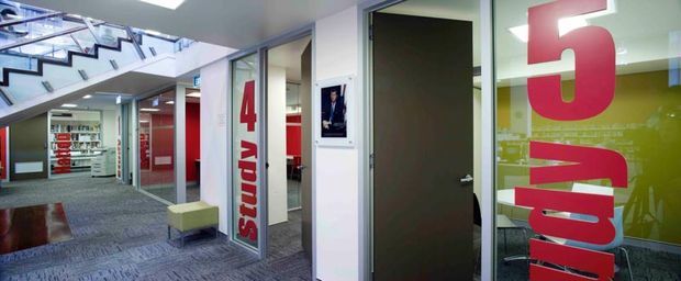 Interior view of a contemporary educational or commercial corridor, with carpeted flooring, glass-partitioned rooms, bold red-and-white study signage, a dark door, and a visible staircase. The space has bright fluorescent lighting and an open, institutional fit-out.
