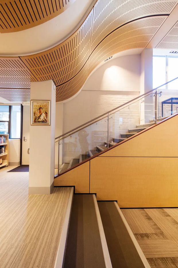 Interior view of a contemporary multi-level commercial or institutional space featuring a sculptural curved timber slatted ceiling, warm wood stair and balustrade panels, glass stair guards with metal handrails, pale painted masonry walls, and carpeted steps and floor. The composition focuses on a stairway and landing with an adjacent open workspace visible in the background.