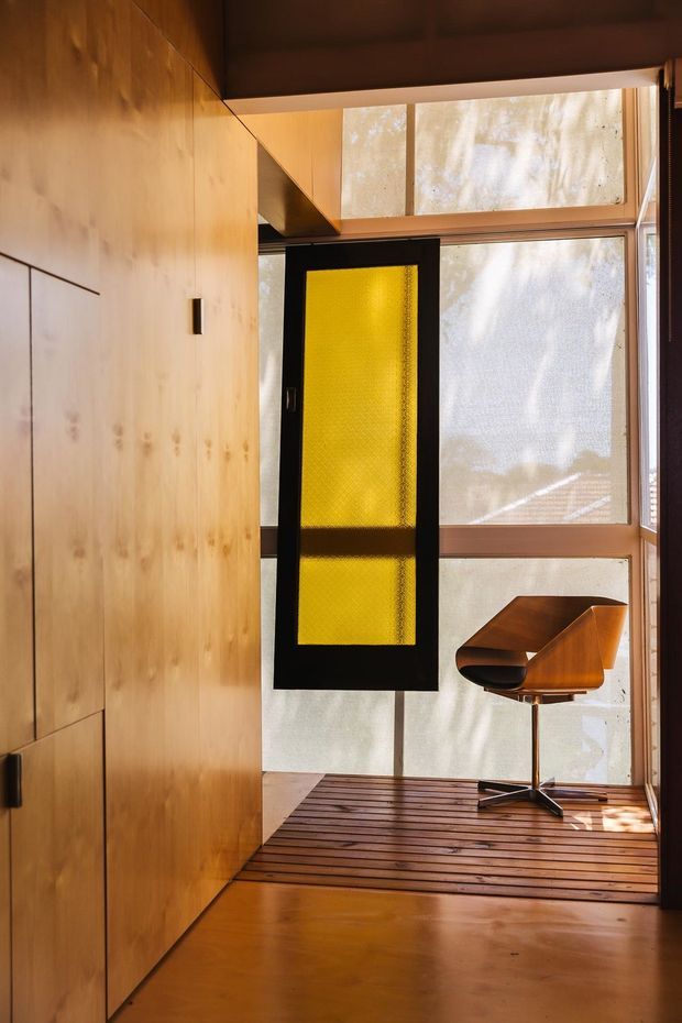 A compact, warm-toned interior with extensive timber wall panelling and built-in cabinetry, a polished timber floor, and large translucent glazed windows. A black-framed pivot or hinged door with yellow translucent glazing creates a strong feature, while a sculptural wooden swivel chair sits beside the window. The space has a refined mid-century character with abundant natural light and a strong connection to the glazing.