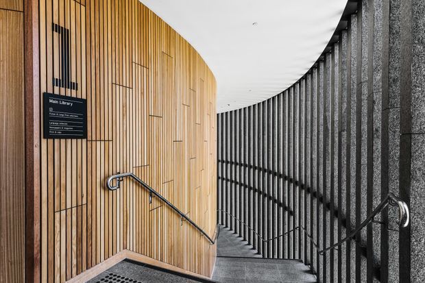Curved stairway descending through a contemporary public interior, with warm vertical timber wall panelling on one side and dark textured stone or concrete wall surfaces with black metal balustrades on the other. A sign identifies the space as the Main Library. The composition emphasizes the sweeping curved ceiling and stair enclosure.