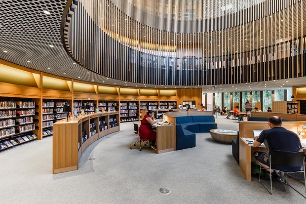 A spacious contemporary library or learning commons with curved timber bookshelves, individual study desks, blue upholstered seating, and a dramatic suspended vertical-slat ceiling feature over a circular central area. The space is brightly lit and arranged for reading, study, and quiet collaboration.