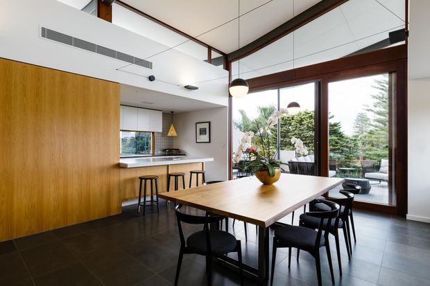 A bright, contemporary open-plan dining and kitchen area with a large timber dining table, black dining chairs, timber cabinetry, a compact island with bar stools, dark tiled flooring, and expansive dark timber-framed glazing opening to a landscaped outdoor setting. A white paneled ceiling, exposed dark timber beams, and pendant lighting add architectural character.
