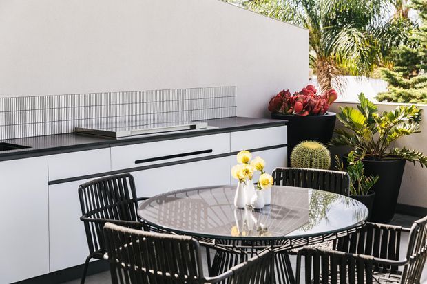 A bright contemporary outdoor dining area with a built-in barbecue kitchen, white cabinetry, dark benchtop, tiled splashback, and a round glass dining table surrounded by black woven chairs. Large potted tropical plants add greenery against a pale rendered wall.
