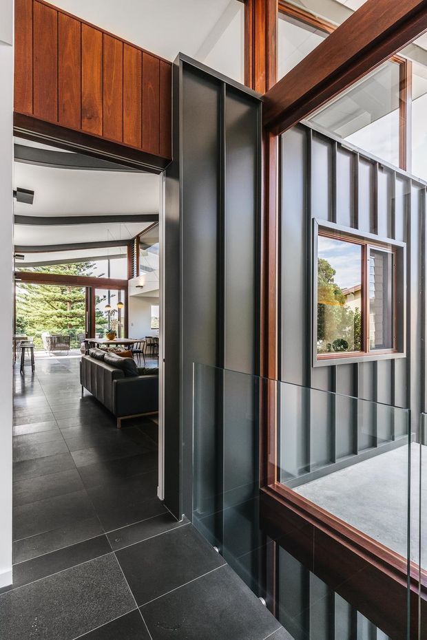 Interior view of a contemporary home showing a dark tiled entry or hallway beside a dramatic glazed stairwell or void. The space features charcoal vertical metal wall panels, rich timber-framed glazing and upper cabinetry, a glass balustrade, and a view through to an open-plan living area with large windows.