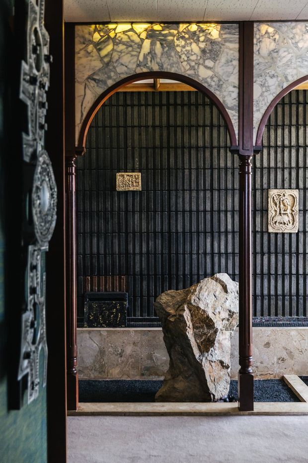 A richly detailed interior feature wall with dark vertically ribbed tiles, pale veined stone panels, and a series of rounded timber arches supported by slender columns. Decorative relief plaques, a large natural rock feature, and dark stone flooring create a dramatic, historic and sculptural atmosphere.