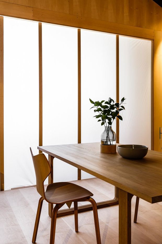 Warm minimalist dining area featuring a solid timber dining table and matching sculptural wooden chair, set against full-height translucent panels framed in timber. The room has pale flooring, extensive timber wall and ceiling surfaces, and restrained natural decor including a potted plant and ceramic bowl.
