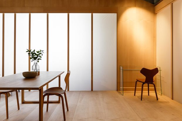 Warm minimalist dining area with a timber dining table and chairs, a small plant centerpiece, pale timber flooring, and extensive translucent or frosted vertical glazing framed in timber. A sculptural dark chair sits in a softly lit timber-lined alcove beside glass shelving.