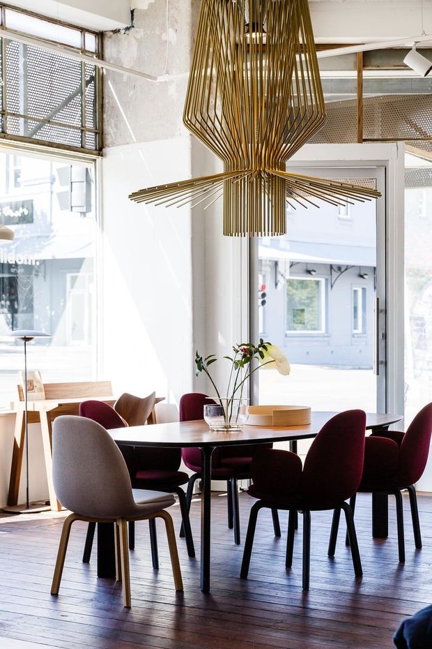 Bright contemporary dining area in a converted commercial-style space, featuring a large round dark dining table, upholstered dining chairs in burgundy, grey and brown, timber flooring, expansive industrial windows and doors, and a dramatic sculptural brass-gold pendant light. Exposed weathered wall surfaces and perforated metal window screens add an industrial character.
