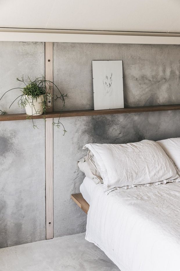 A close-up view of a minimalist bedroom with a low platform bed dressed in wrinkled white linen bedding. The room features mottled grey concrete-look wall panels, pale vertical timber battens, a dark timber picture ledge, and a hanging plant beside a simple monochrome artwork. The palette is restrained and neutral with a raw, tactile material quality.