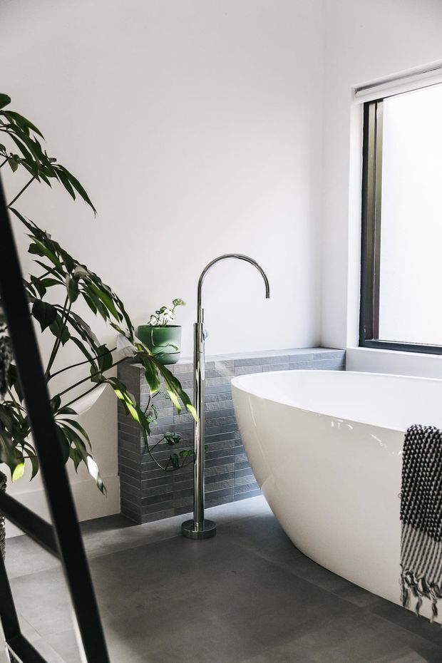 Bright contemporary bathroom featuring a freestanding white bathtub, floor-mounted chrome bath filler, dark grey tiled and concrete-look surfaces, a black-framed window, and lush indoor greenery. The space has a clean, minimalist aesthetic with abundant natural light.