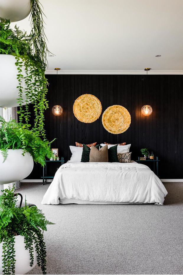 A bright, contemporary bedroom centred on a neatly made bed with white linen, layered neutral and dark green cushions, and black vertical wall panelling behind the headboard. Two woven circular wall artworks provide a warm natural accent, while globe pendant lights flank the bed. Large cascading potted plants frame the left side of the room, and the floor is covered in light grey carpet.