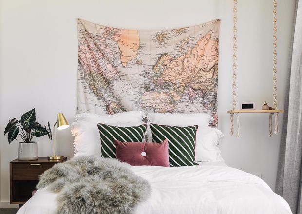 A styled bedroom with a white bed, layered pillows, a green-and-white striped accent cushion, and a mauve lumbar pillow. A large vintage-style world map tapestry hangs above the bed, while a dark timber bedside table, brass lamp, potted plant, small wall shelf, and grey curtain complete the room. The palette is predominantly white with green, pink, and natural timber accents.