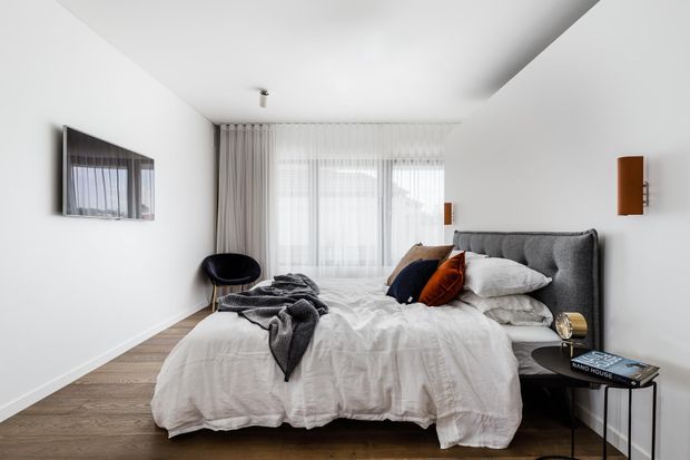 A bright, contemporary bedroom with a large upholstered grey bed, white textured bedding, dark accent cushions, timber flooring, sheer curtains, and minimalist bedside furniture. The room has white walls, a recessed ceiling detail, and a framed wall artwork.