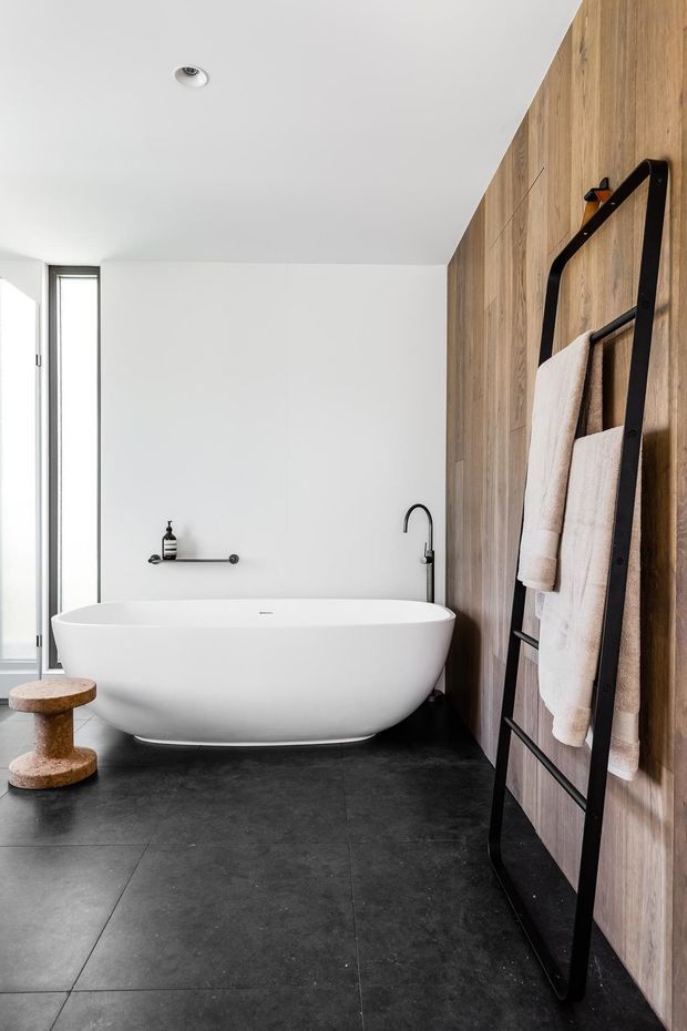 A contemporary bathroom featuring a freestanding white bathtub, matte-black floor and wall-mounted fixtures, dark large-format floor tiles, and a warm timber wall with integrated black towel ladder. A narrow vertical window provides natural light, and a small wooden stool sits beside the bath.