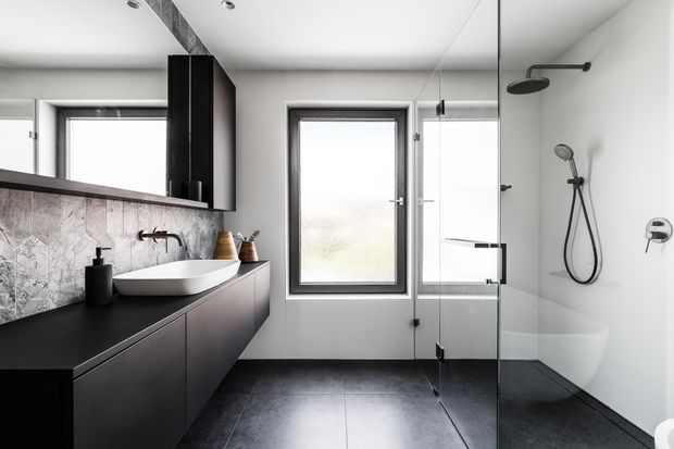 Contemporary bathroom with a wall-mounted dark vanity, white vessel basin, large mirrored cabinetry, grey stone-look splashback, freestanding bath, and a glass-enclosed shower. The room has dark grey floor tiles, white walls, black fixtures, and a window providing natural light.