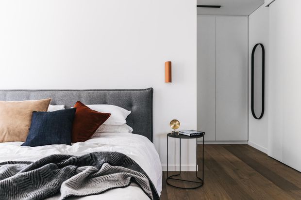 A contemporary bedroom with a low-profile upholstered grey headboard, layered white bedding, and contrasting navy, tan, and rust cushions. A slim black metal side table, wall-mounted cylindrical light, full-height pale built-in wardrobes, dark timber flooring, and a tall rounded mirror create a clean, refined interior.