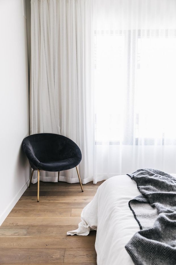 A bright, minimalist bedroom corner with a partially visible bed dressed in white bedding and a dark textured throw. A black upholstered accent chair with slim brass-toned legs sits beside full-height sheer and heavier white curtains. The room features pale timber flooring, white walls, and abundant natural light.