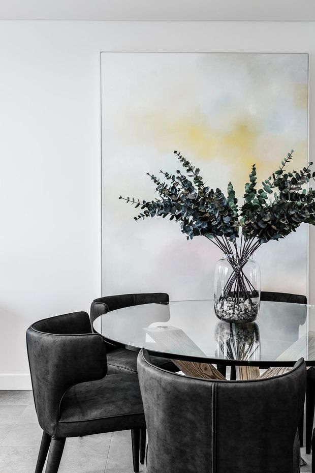 Refined dining area with a round glass dining table, sculptural dark upholstered dining chairs, and a large transparent vase filled with eucalyptus branches. A soft abstract artwork provides a subtle neutral and pastel backdrop against white walls, creating a sophisticated monochromatic interior.