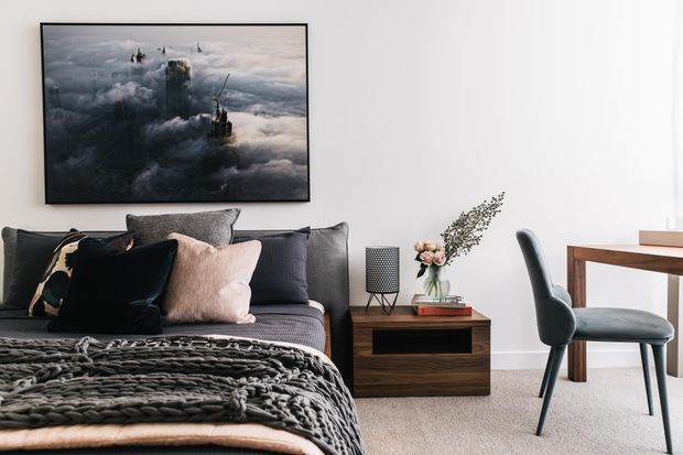 A contemporary bedroom with a low upholstered bed, layered grey, black and blush cushions, and a chunky charcoal knitted throw. A dark wood bedside table with decorative objects sits beneath a dramatic framed artwork, while a simple wood desk and upholstered chair occupy the corner. The room has white walls, pale carpet and a restrained, sophisticated palette.