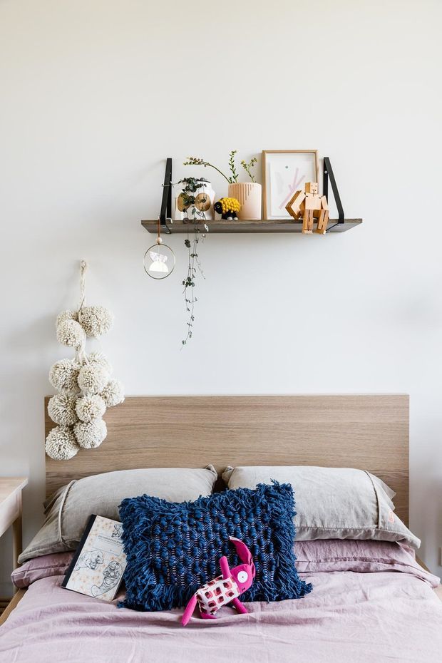 A softly styled bedroom with a light timber bedhead, pale bedding, and layered grey and mauve pillows. A dark blue textured cushion and bright pink decorative toy add colour. Above the bed is a simple timber display shelf with black brackets, framed artwork, small ornaments, plants, and hanging decor. The room has white walls and a calm, playful feel.