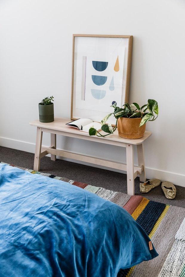 A bright, softly styled bedroom corner featuring a light timber console table against a white wall, a framed abstract print, and several potted plants. The foreground shows part of a bed with a vivid blue coverlet and a multicoloured woven rug over dark carpet. Woven slippers sit beside the console.