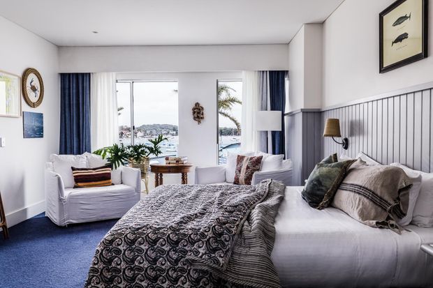 A bright coastal-style bedroom with a large bed dressed in layered white and patterned bedding, a matching upholstered armchair, blue carpet, navy-and-white curtains, and grey-blue vertical wall panelling behind the bed. Large windows overlook a waterfront or harbour setting, while framed artwork, a wall-mounted reading lamp, plants, and a small round side table add character.