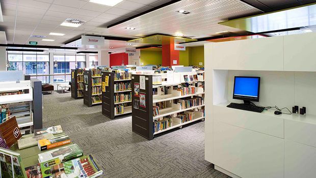 Bright contemporary library interior with rows of bookshelves, reading and computer stations, colourful wall sections, patterned carpet, and a suspended acoustic ceiling. The space appears to be a public or educational library, with signage and books arranged throughout.