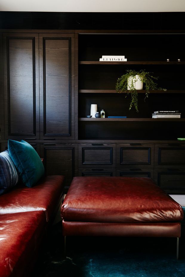 A moody, richly layered living room detail featuring dark timber built-in cabinetry with recessed panel doors and open display shelving. Deep burgundy leather seating and ottoman sit over a dark teal rug, with teal and blue cushions adding contrast. Minimal decor, books, pottery, and a trailing plant are arranged on the shelves.