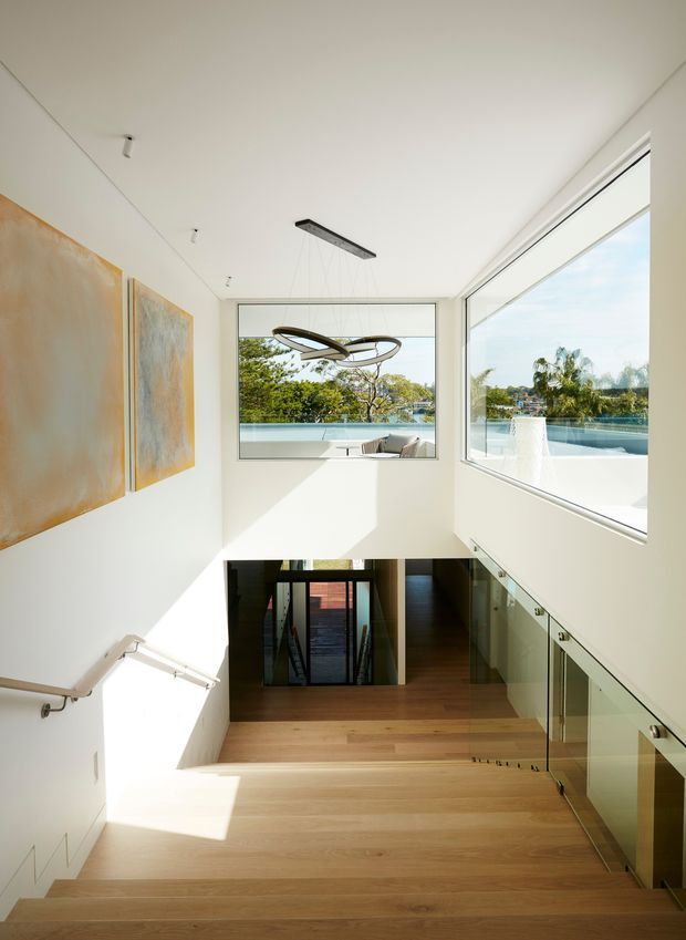 Bright contemporary stairway and landing overlooking a lower-level entry or living area. The space features pale timber flooring, white walls and ceiling, frameless glass balustrades, expansive corner glazing, recessed spotlights, abstract wall art, and a sculptural suspended pendant. Trees, water, and an outdoor pool are visible beyond the windows.