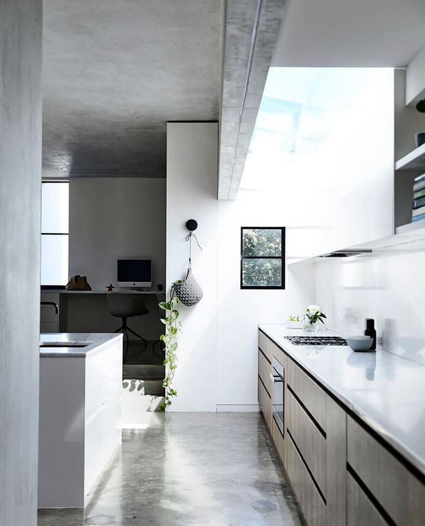 Bright contemporary kitchen with a long white benchtop, integrated cabinetry, a central island, polished concrete flooring, and a raw concrete ceiling beam. A compact home office is visible beyond the kitchen, with abundant natural light and indoor greenery.
