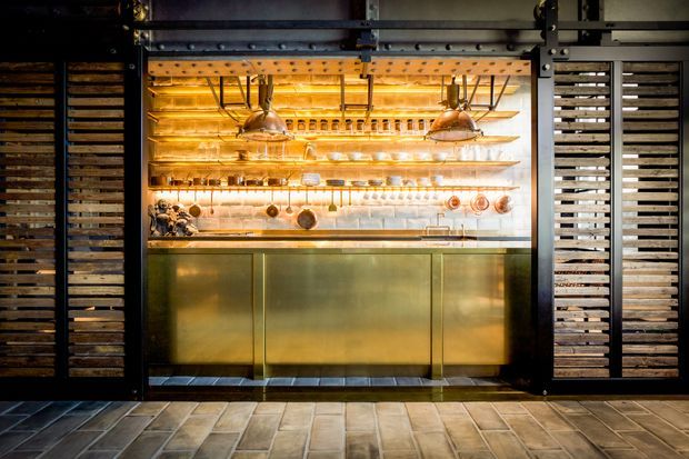 A moody commercial bar or service counter framed by dark industrial timber screens. The illuminated back bar has brass-toned cabinetry, open shelving, hanging glassware, copper cookware, bottles, and warm pendant lights. Large-format rectangular tile flooring and exposed dark metal overhead hardware reinforce the industrial hospitality setting.