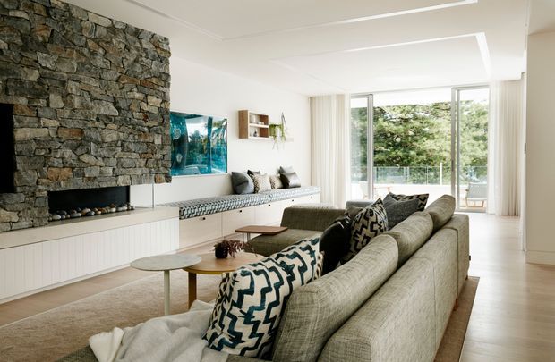 Bright contemporary living room with a large textured stacked-stone fireplace wall, integrated white built-in cabinetry and bench seating, expansive glazed doors, pale timber flooring, and a large neutral sectional sofa with patterned cushions. The room opens to a leafy outdoor setting and has a refined, relaxed character.