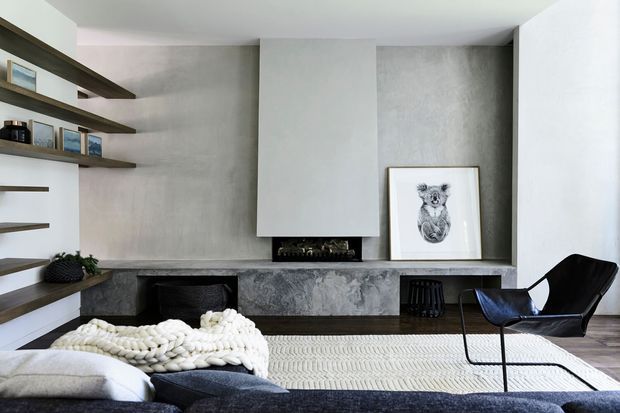 Contemporary living room with a restrained grey and white palette, textured concrete-look fireplace surround, floating dark timber shelving, low built-in joinery, and a sculptural black lounge chair. A large sectional sofa, chunky knitted throw, and patterned rug create a soft contrast against the minimalist built-in architecture.
