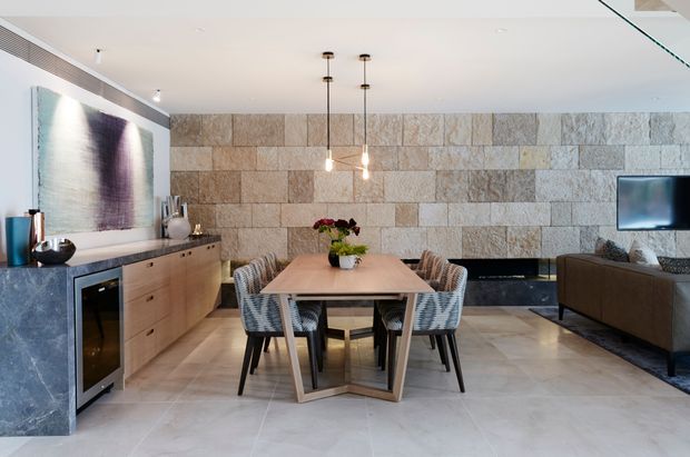A refined open-plan dining and living area with a large rectangular dining table and upholstered chairs positioned centrally. The room features pale stone-look tiled flooring, a dramatic full-height wall clad in rectangular stacked stone tiles, a dark stone kitchen or bar counter at left with integrated appliances, a timber sideboard, abstract wall art, pendant lighting, and a television over a low fireplace or media wall.