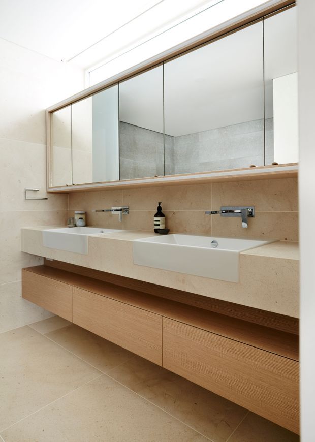 Bright contemporary bathroom vanity with two integrated rectangular basins, wall-mounted chrome taps, a full-width mirrored cabinet, beige stone or porcelain wall and floor finishes, and extensive light timber floating cabinetry.