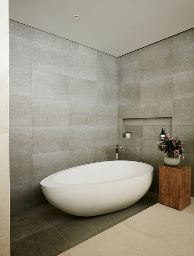 A refined contemporary bathroom featuring a freestanding white oval bathtub against large-format grey stone-look wall tiles. Darker grey floor tiles define the bathing area, while a pale stone or tile floor extends in the foreground. A recessed wall niche contains bath products, and a timber pedestal with a floral arrangement adds warmth.