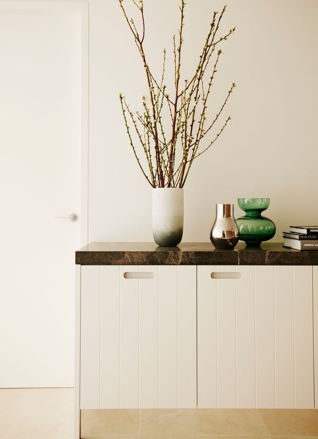 A refined minimalist interior vignette featuring a long white built-in cabinet with vertically grooved doors and recessed handles, topped by a dark veined stone surface. Decorative ceramic and metallic vessels, stacked books, and tall bare branches add styling. A plain white door and warm pale tiled floor reinforce the clean, neutral scheme.