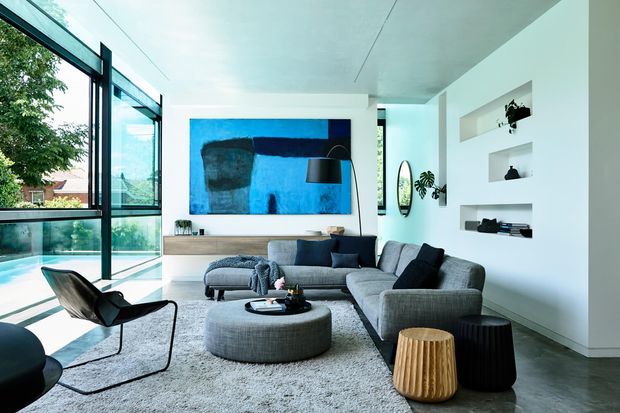 Bright contemporary living room with floor-to-ceiling black-framed glazing, a large grey sectional sofa, round ottoman, sculptural lounge chair, built-in white display niches, timber console joinery, and a bold blue abstract artwork. The room opens toward a glazed outdoor area and is finished with a polished grey floor and light rug.