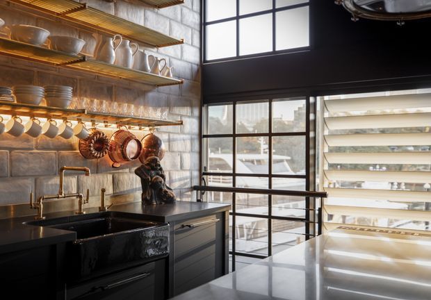 A dark, sophisticated industrial-style kitchen with open timber shelves displaying crockery and glassware, a tiled or block-textured feature wall, copper cookware hanging above a deep black sink, dark built-in cabinetry, and a large stone island or benchtop. Black-framed industrial glazing brings in strong daylight and overlooks neighbouring buildings.