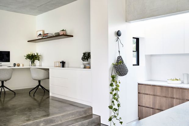 Bright open-plan interior combining a compact home office and kitchen. The space features white walls and cabinetry, a polished concrete ceiling and stepped concrete floor, a dark timber floating shelf, wood-grain kitchen cabinetry, integrated white overhead cupboards, and minimalist furnishings with indoor plants.