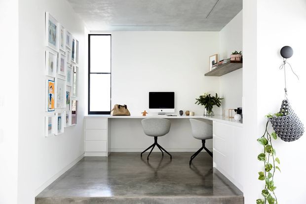 Bright compact home office with a built-in desk spanning the back wall, two modern stools, integrated white drawers and cabinetry, a computer workstation, floating shelf, indoor plants, framed artwork, and a polished grey concrete floor. The room has a clean minimalist fit-out and a tall narrow black-framed window.