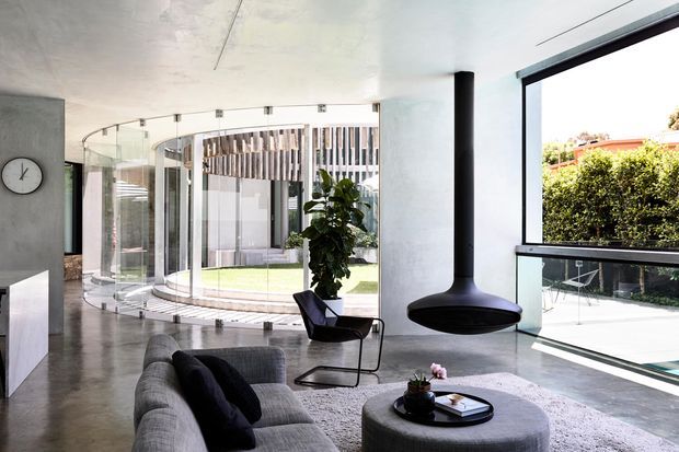 Bright contemporary living room with polished concrete flooring, a large grey sectional sofa, rounded ottoman coffee table, sculptural black suspended fireplace, and expansive glazing. A curved glass wall opens visually toward a landscaped courtyard, while a minimalist kitchen or joinery element and wall clock sit at the left.