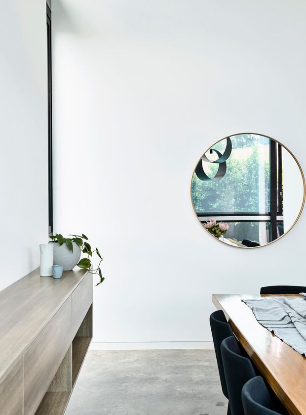 Bright minimalist dining area with a long timber dining table and dark upholstered chairs, positioned beside a pale timber built-in sideboard. White walls, polished concrete flooring, a large round framed mirror, and simple indoor plants create a clean contemporary interior. The mirror reflects glazing and greenery outside.