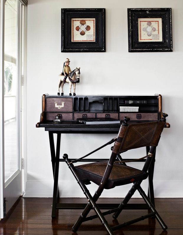 A compact, carefully styled writing nook beside a glazed door, featuring a dark vintage secretary desk with leather-look compartments and drawers, a woven leather campaign-style folding chair, small equestrian figurine, and two framed artworks on a light wall. Dark timber flooring and black furniture create a refined, classic mood.