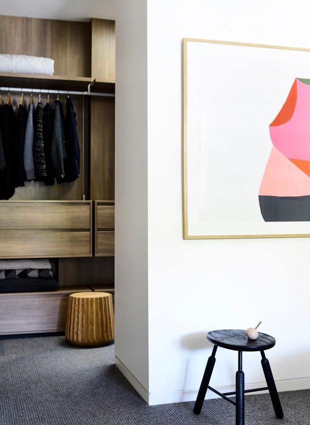A refined bedroom dressing area with an open built-in wardrobe featuring warm timber joinery, hanging clothing, drawers, and shelving. A white wall displays a framed abstract artwork, while small sculptural stools sit on a grey carpet. The space has a clean, contemporary feel with warm wood accents and restrained furnishings.