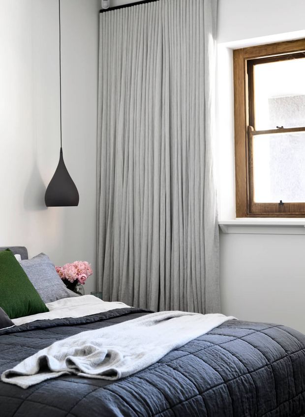 A close-up of a modern bedroom featuring a neatly made bed with a dark grey quilted coverlet and soft white throw, layered grey and green pillows, full-length pleated grey curtains, a minimalist black pendant light, and a timber-framed window. The room has a calm, refined palette of white, grey, charcoal, green, and natural timber.